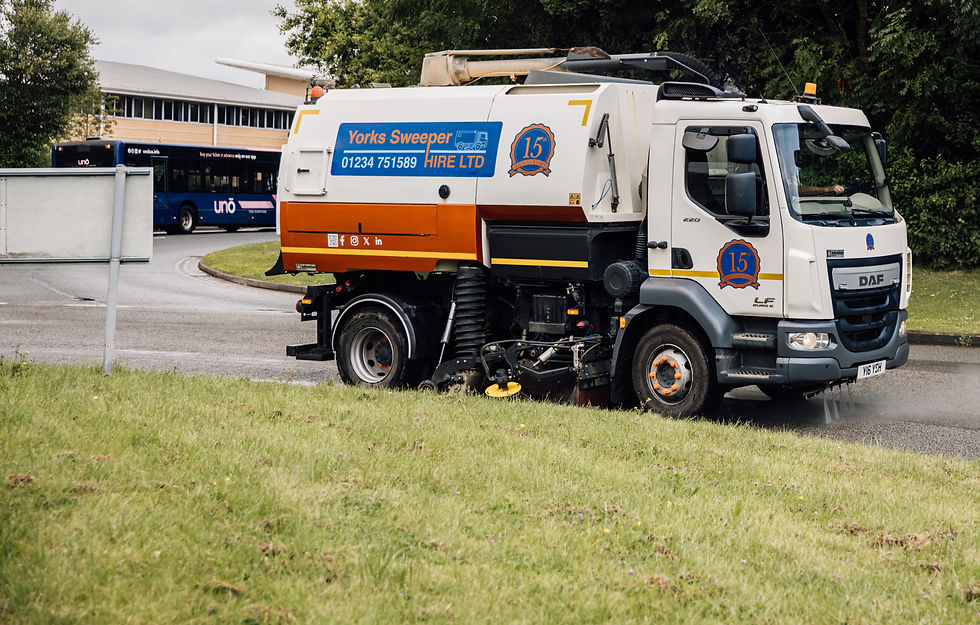 Road sweeper truck in motion sprays water on the road, "Yorks Sweeper Hire LTD" logo visible. Background shows greenery and a bus.