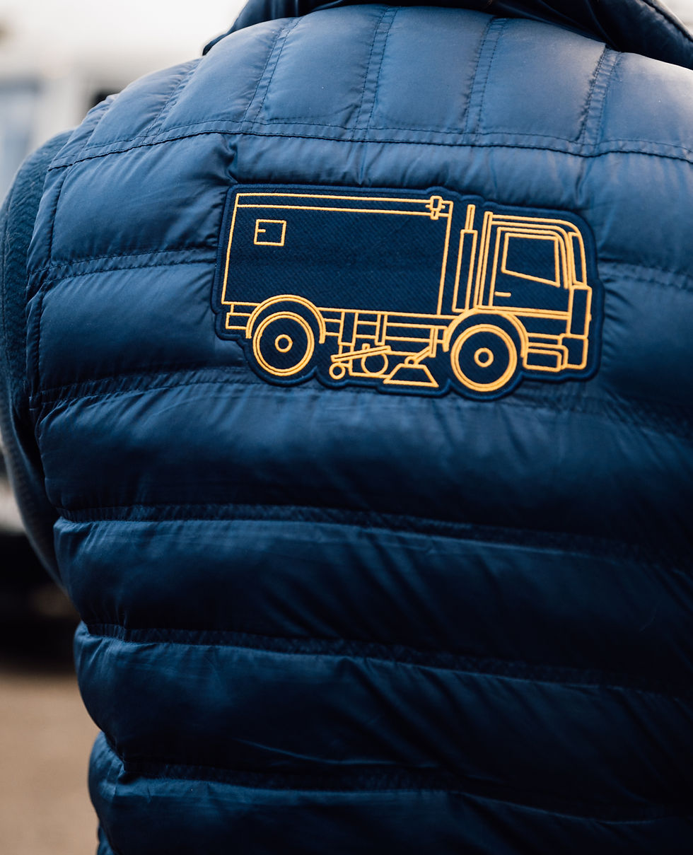 Close-up of a blue jacket with a yellow Road Sweeper truck design on the back, seen outdoors. The image has a casual and rugged feel.
