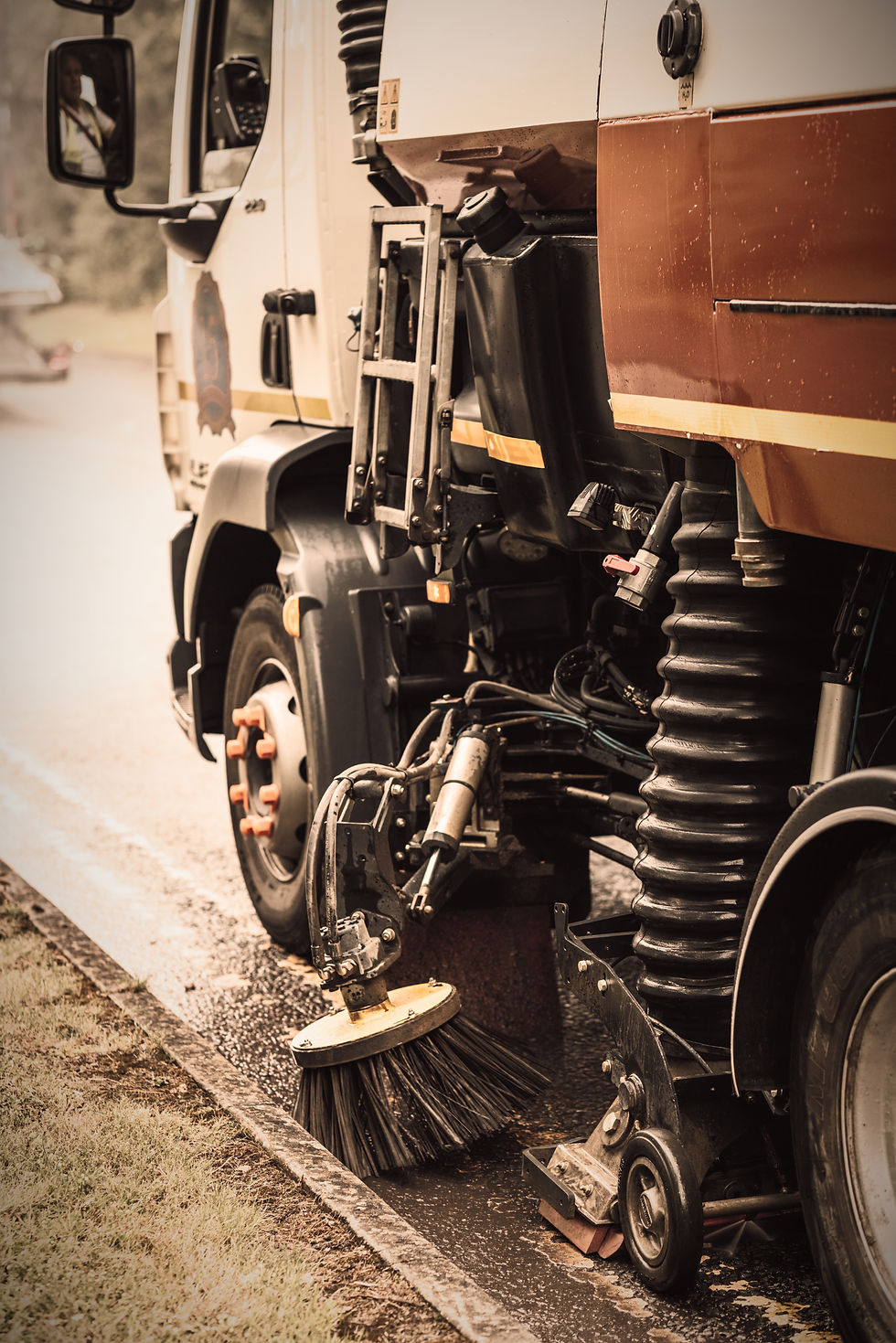 Street sweeper truck with rotating brush cleans wet road. Sepia tones create a vintage feel. Grass visible on roadside.