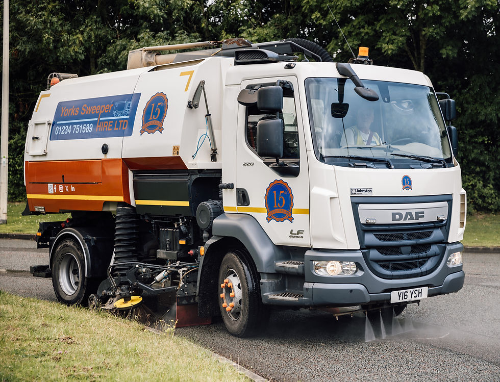 Street cleaning truck with orange and white design sprays water on the road. Trees in the background. "Yorks Sweeper Hire LTD" text visible. Road Sweeper.