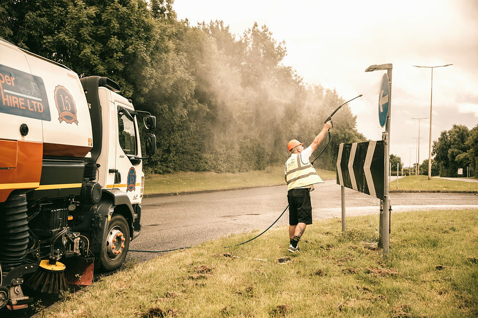 Worker in a hi-vis vest and hard hat uses a hose to clean a road sign next to a road cleaning truck. Trees line the road in the background.