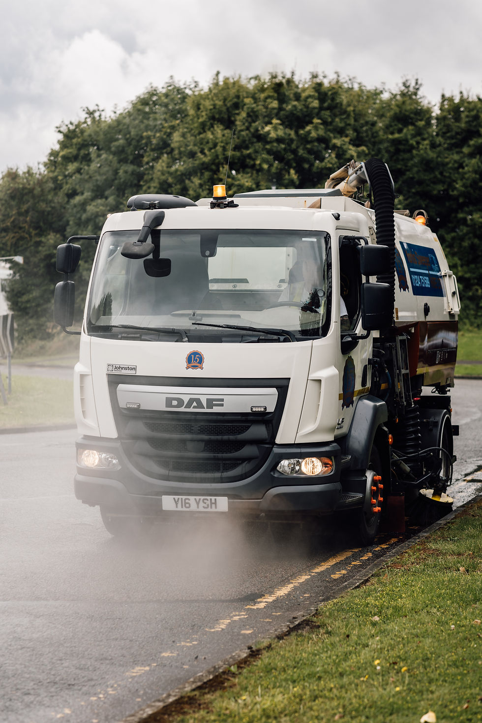 White Road sweeper truck cleaning a road with water spray on a cloudy day, surrounded by green trees and grass. License plate: Y16 YSH.