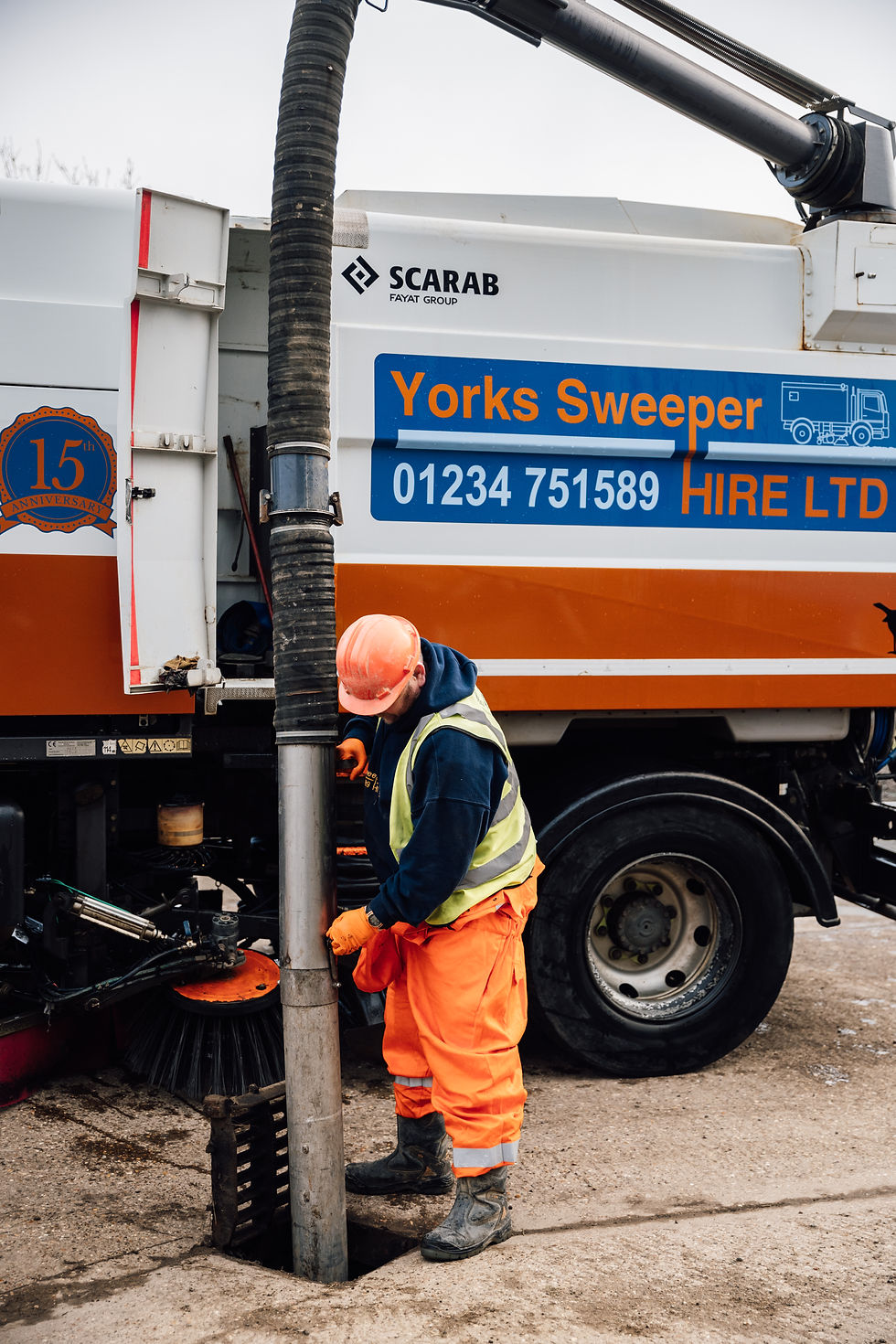 Worker in orange gear operates a vacuum hose from a street sweeper truck. "Yorks Sweeper HIRE LTD" sign visible. Industrial setting. Gully Sucker. Drain clearing, drain cleaning.