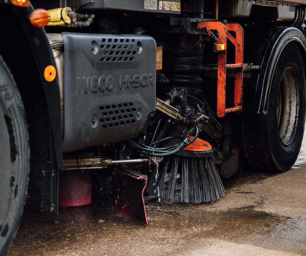 Road sweeper truck cleaning wet pavement; close-up of rotating brushes and equipment. Gray, black, and red details with visible water.