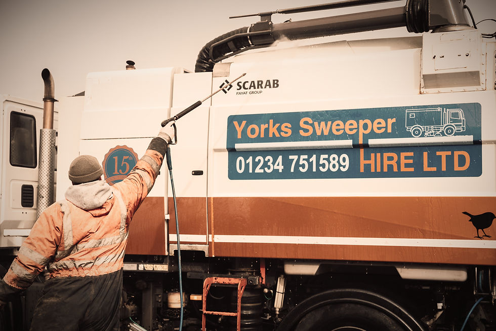 Worker in orange gear pressure washes a street sweeper with "Yorks Sweeper Hire LTD" info visible on the truck. Overcast day.