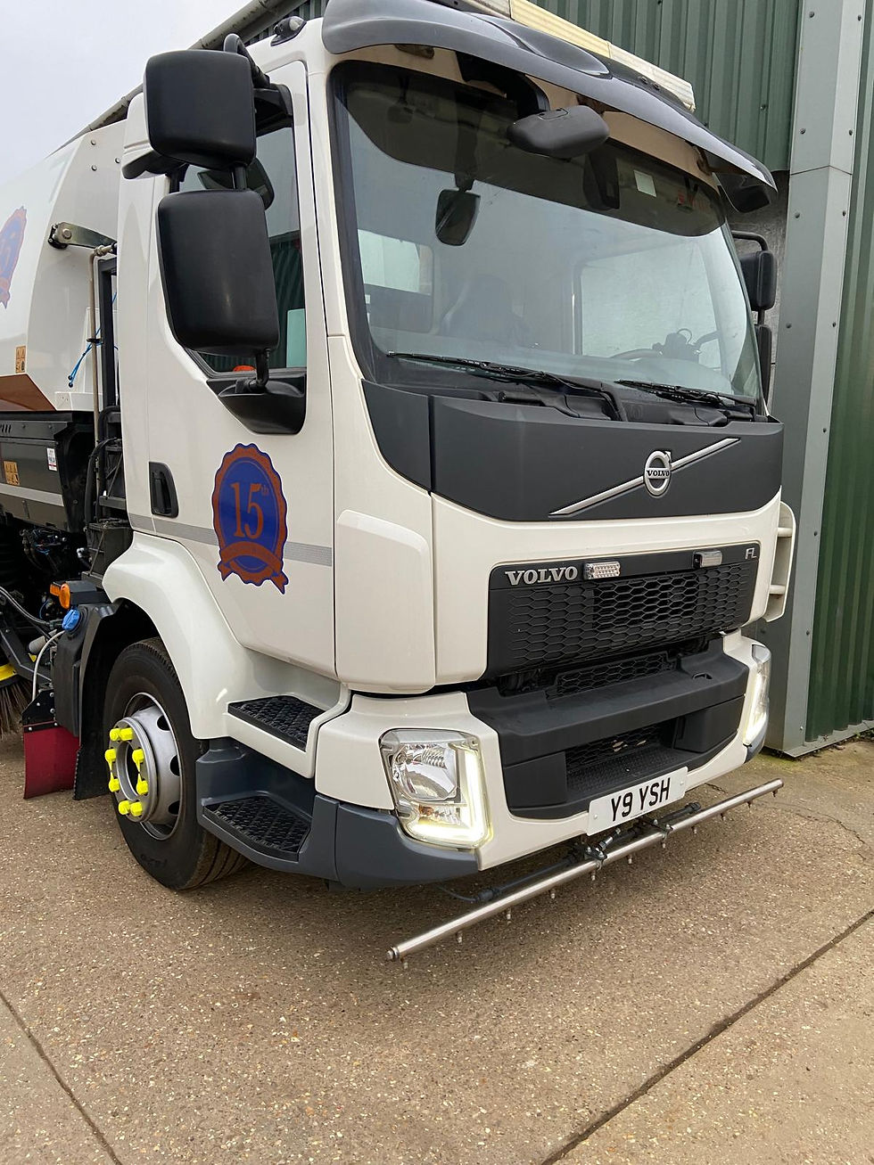 White Volvo Road Sweeper truck parked on concrete near a green building, featuring a "15" emblem and plate "Y9 YSH" with a visible cleaning mechanism. Yorks Sweeper Hire