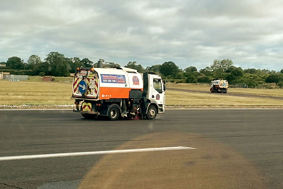 Street sweeper on a tarmac road, with another in the distance, against a green field and cloudy sky. "Yorks Group" text visible on truck.