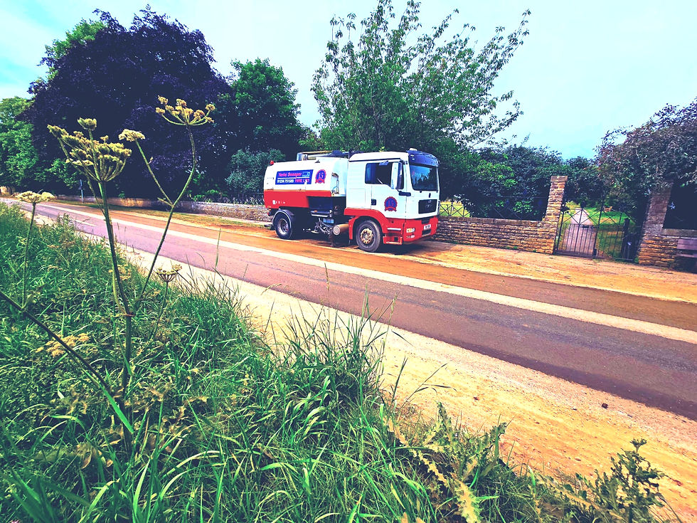 Road Sweepers Oxfordshire. A red and white truck drives down a rural road, bordered by green grass and trees. A stone wall and iron gate are in the background.
