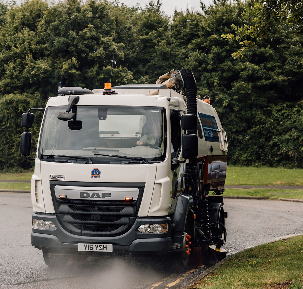 Road sweeper truck cleaning a road near a park with green trees in the background. The truck is white with "DAF" and license plate "Y16 YSH".