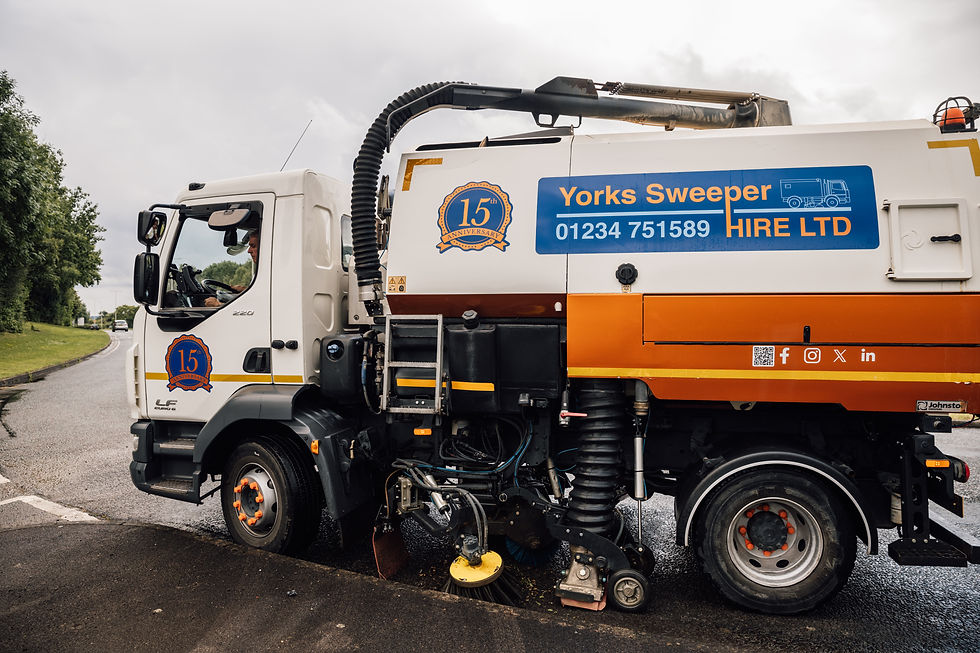 Road sweeper truck with "Yorks Sweeper Hire LTD" and contact info, on a suburban road. Overcast sky, vehicle marked with anniversary badge.