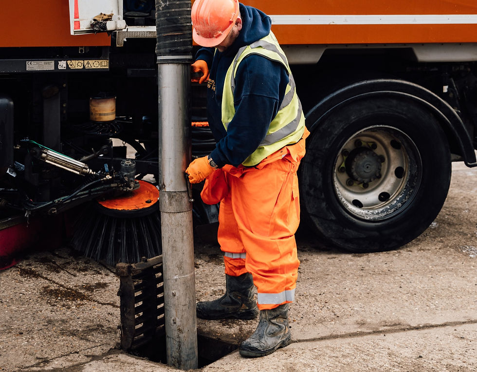 Worker in orange safety gear operates street sweeper truck. Gray tube and brush visible. Industrial setting, overcast mood. Road Sweeper, Gully Sucker, Drain Clearing. Yorks Sweeper Hire Ltd.