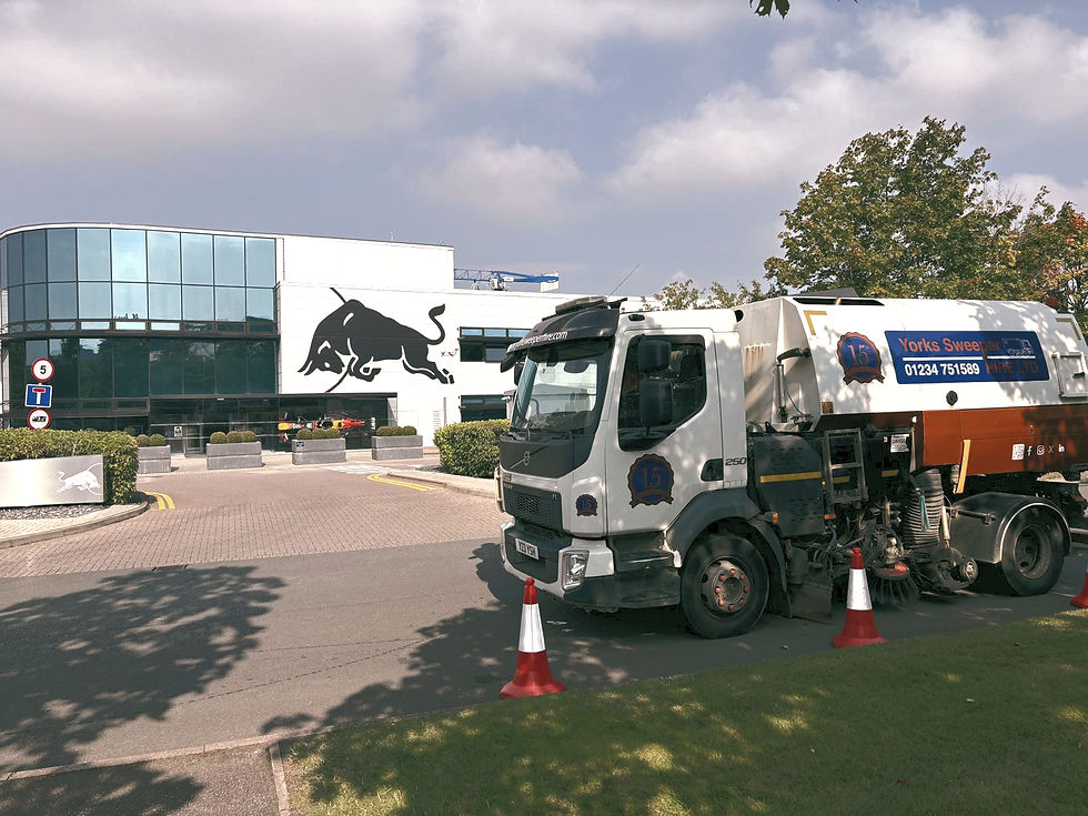Street sweeper truck parked outside a modern building with large bull logo. Green landscaping, red cones, and vehicle signage visible.