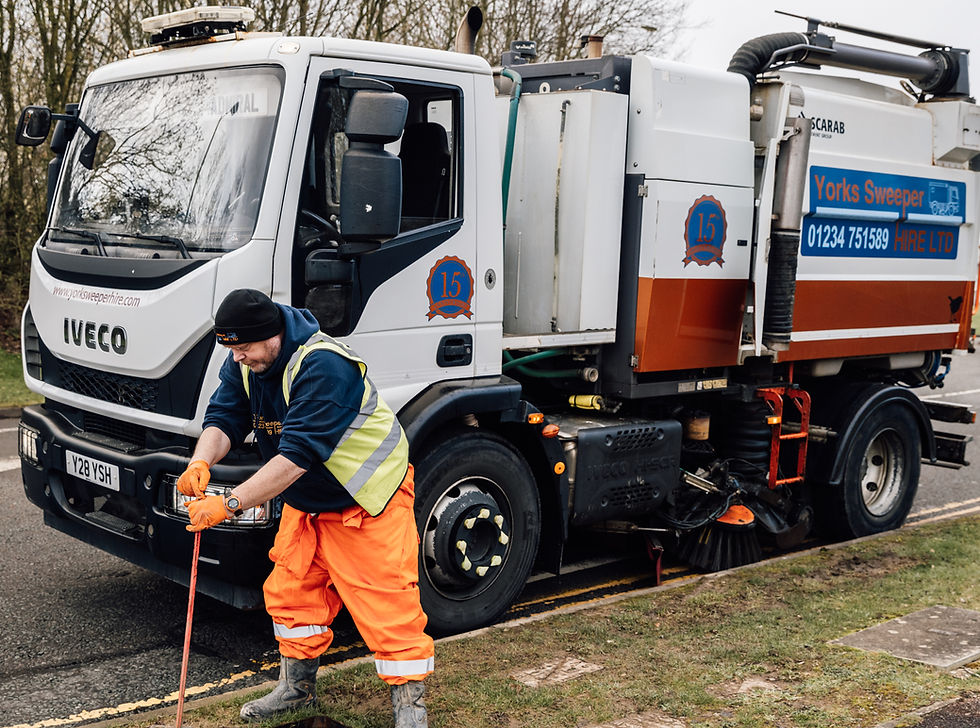 Worker in orange pants and vest uses a tool near a street sweeper truck with "Iveco" and "Yorks Sweeper Hire LTD" branding. Roadside setting.