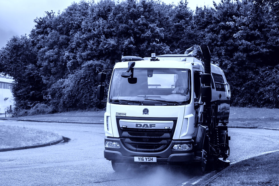 Street sweeper truck cleaning road, spraying water with forest background. The truck is white with visible text "Johnston" and "DAF." Yorks Sweeper Hire