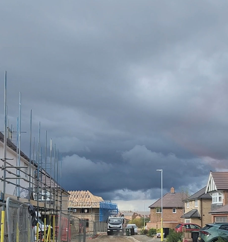 Overcast sky over a suburban street with ongoing construction. Scaffolding, a road sweeper truck, and two houses in view. Dark clouds forecast rain.