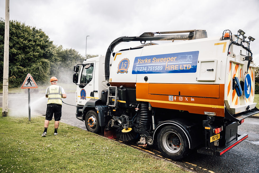Worker in hi-vis vest and orange helmet power-washes a road sign near a street sweeper truck on a cloudy day. "Yorks Sweeper Hire Ltd" sign visible.
