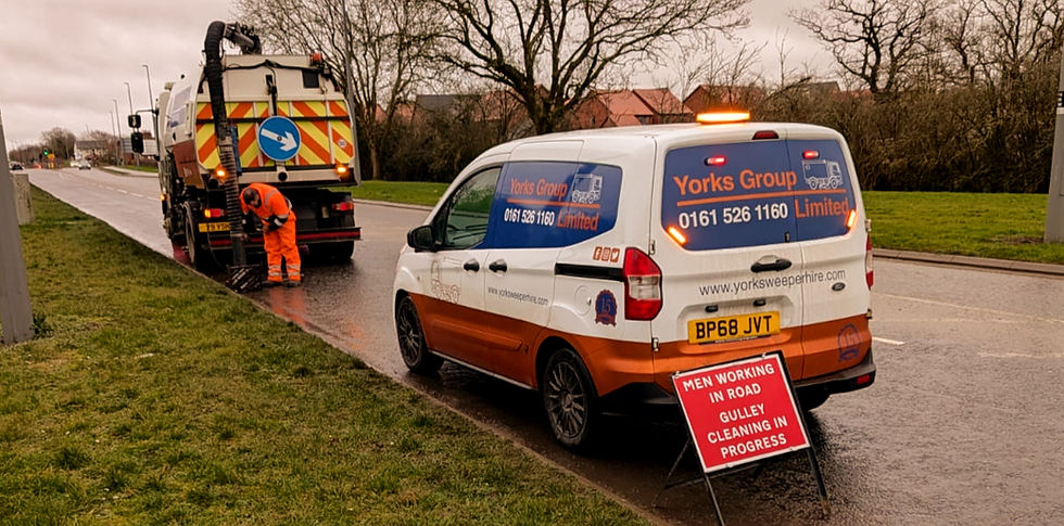 Worker in orange gear cleans gullies beside a road. Two vans labeled Yorks Group are present, with a sign warning of roadwork. Overcast sky.