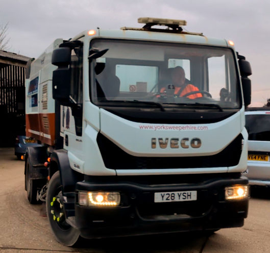 Street sweeper truck labeled "IVECO" with a visible driver in an orange vest. It's parked near another vehicle on a cloudy day.