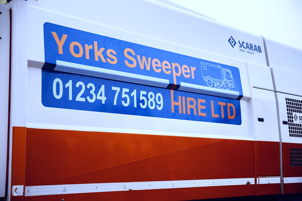 Close-up of a road sweeper truck with text "Yorks Sweeper HIRE LTD 01234 751589". Blue, white, and orange colors; Scarab logo visible.