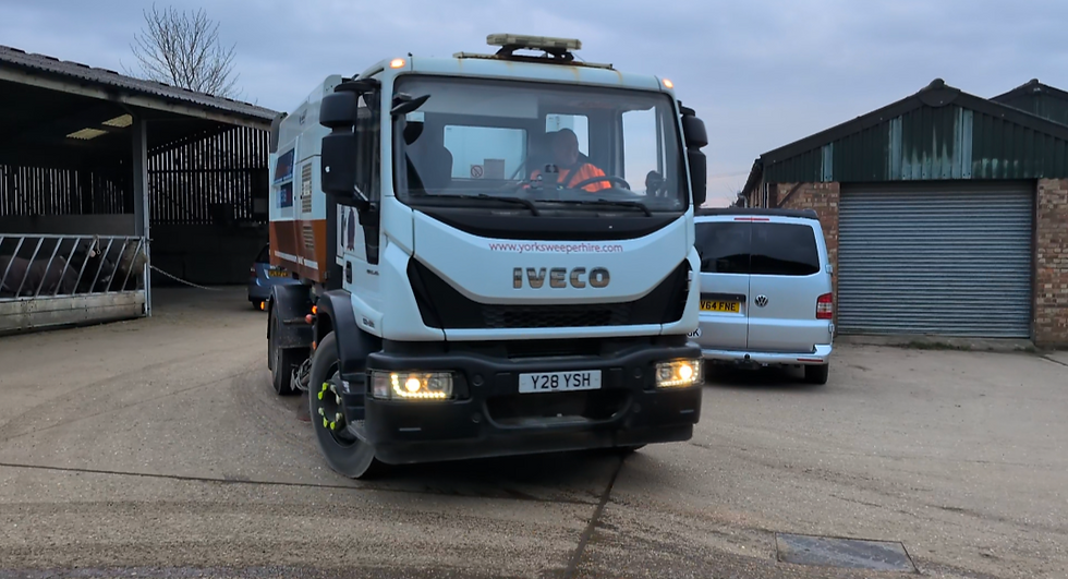 A street sweeper truck with "Iveco" and "www.yorksweeperhire.com" drives on a farmyard. Overcast sky; a van is visible in the background.