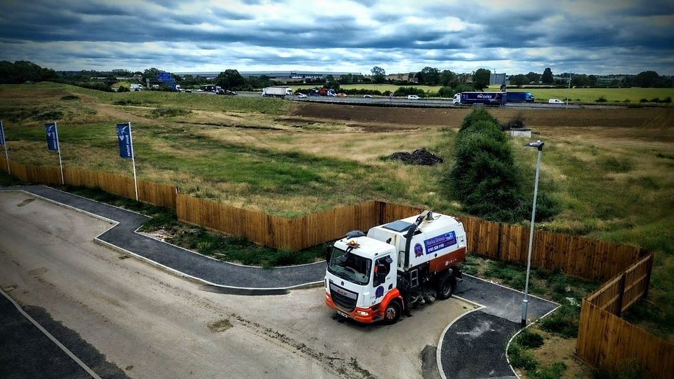 Street sweeper on a paved road next to a wooden fence, with fields and a highway in the background under a cloudy sky. Blue flags line the road.