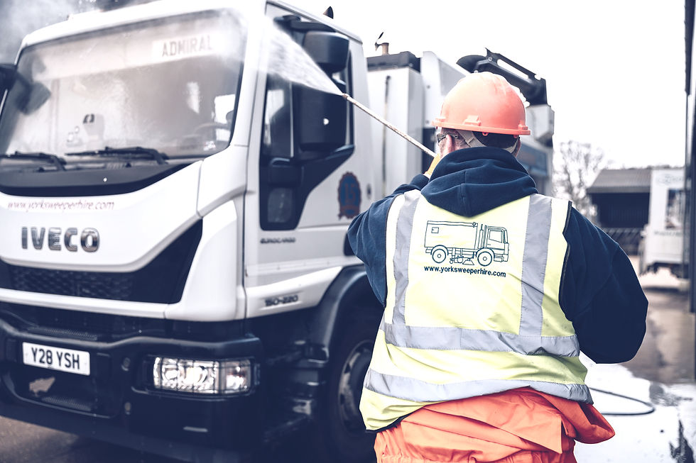 Worker in hi-vis vest and orange helmet pressure washes white "IVECO" Road Sweeper truck labeled "Admiral" outdoors. Overcast sky.
