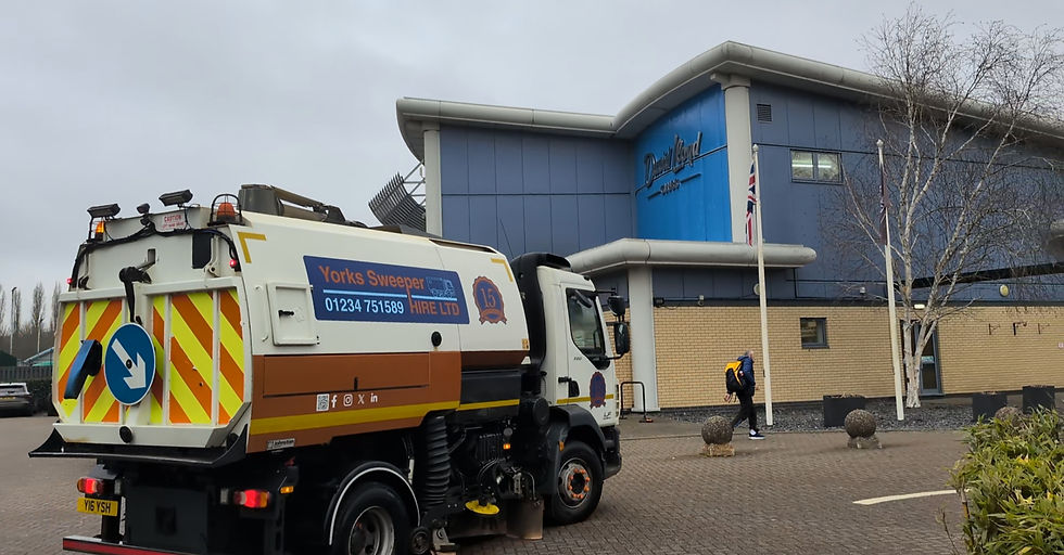 Yorks Sweeper Hire road sweeper truck outside a blue industrial building labeled "David Lloyd." A person with a backpack walks past. Cloudy sky.