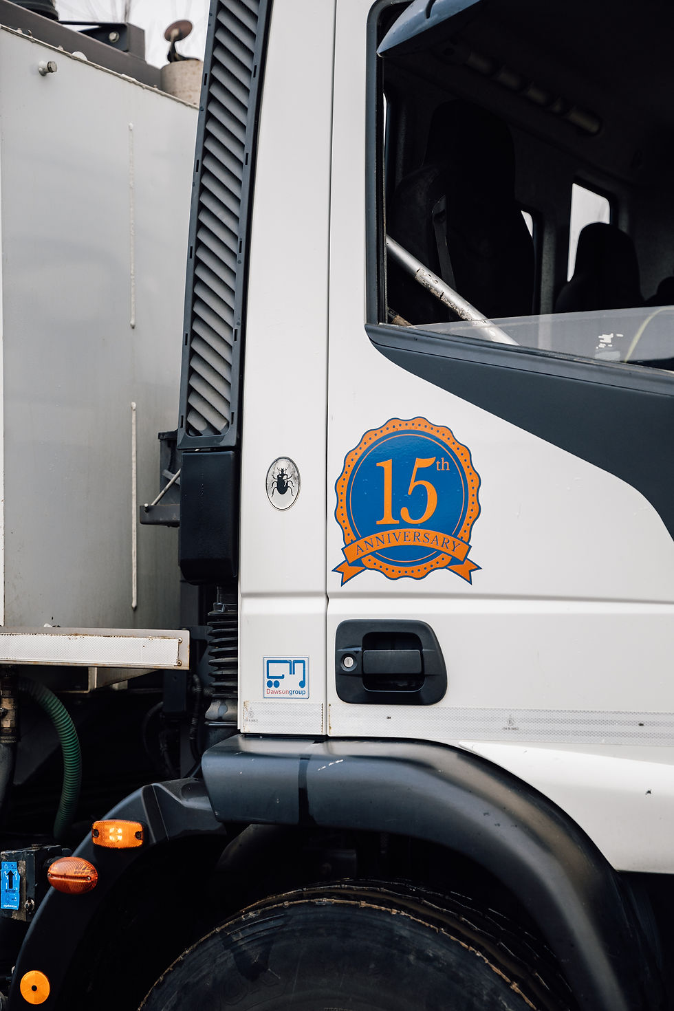 Truck side with "15th Anniversary" badge in blue and orange. Black and white exterior, visible tire, and vehicle details. Urban setting.