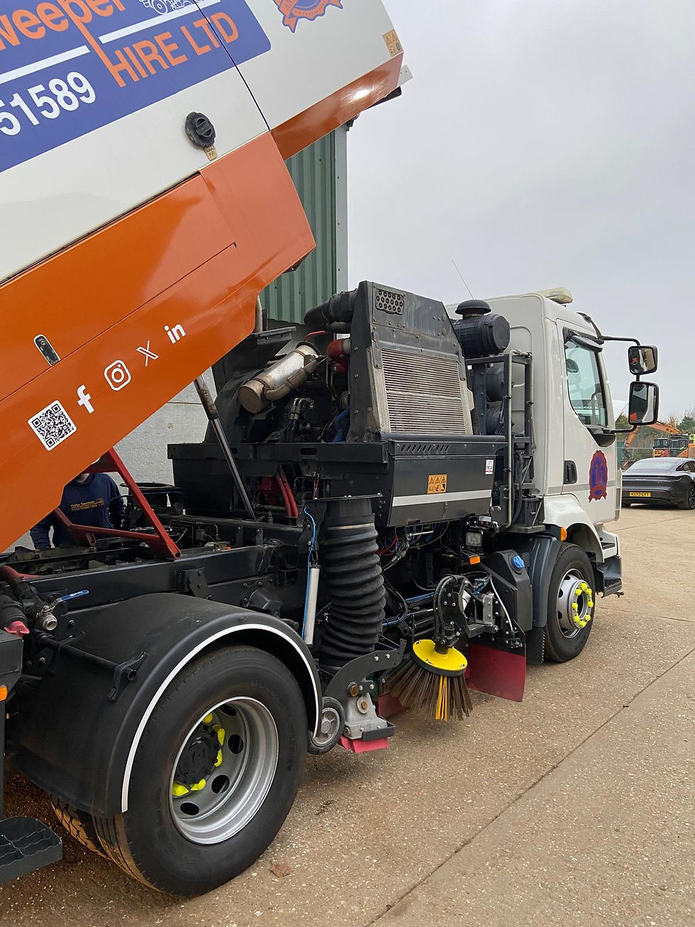 Street sweeper with orange and white signage, showing cleaning brushes and machinery. Overcast sky, building, and car in background.