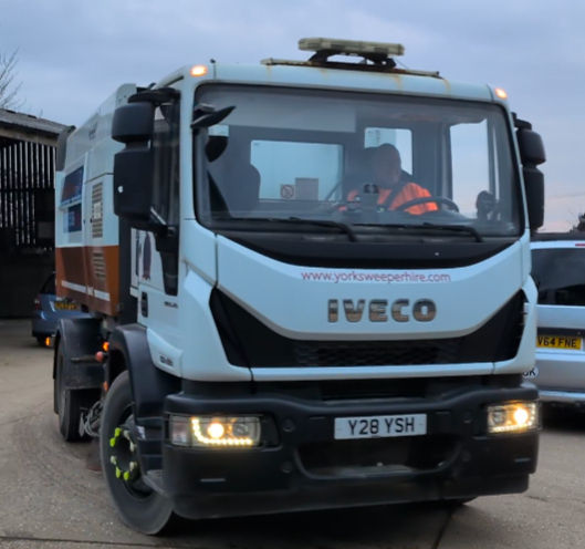 Street sweeper truck with "Iveco" logo, front view. Driver in orange vest. Overcast sky. Website "yorksweeperhire.com" visible.