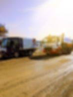 Two sweeper trucks on a muddy construction site. Sunlit sky, scaffolding on the left. Trucks have logos and contact info visible.