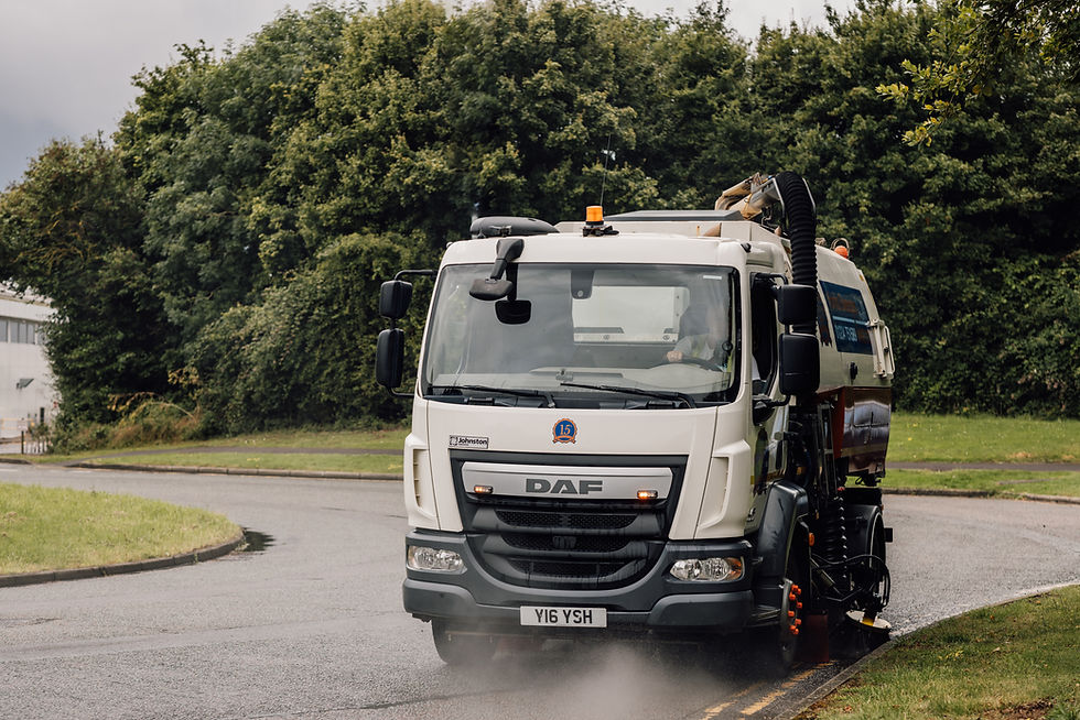 Road sweeper truck cleaning a curved road surrounded by greenery. White truck with visible "DAF" and "Y16 YSH" plate, spraying water.