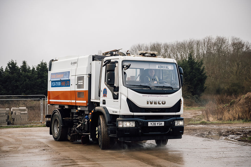 Street sweeping truck labeled on a muddy road, surrounded by trees. Visible text: www.york sweeperhire.com. Overcast sky. Road Sweeper.