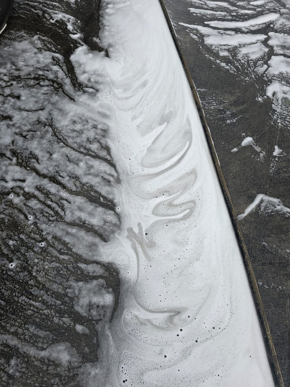 Close-up of swirling white foam on a dark, textured surface, possibly concrete. The image conveys a sense of fluidity and motion. Yorks Sweeper Hire Limited. Chemical Oil Clear up.