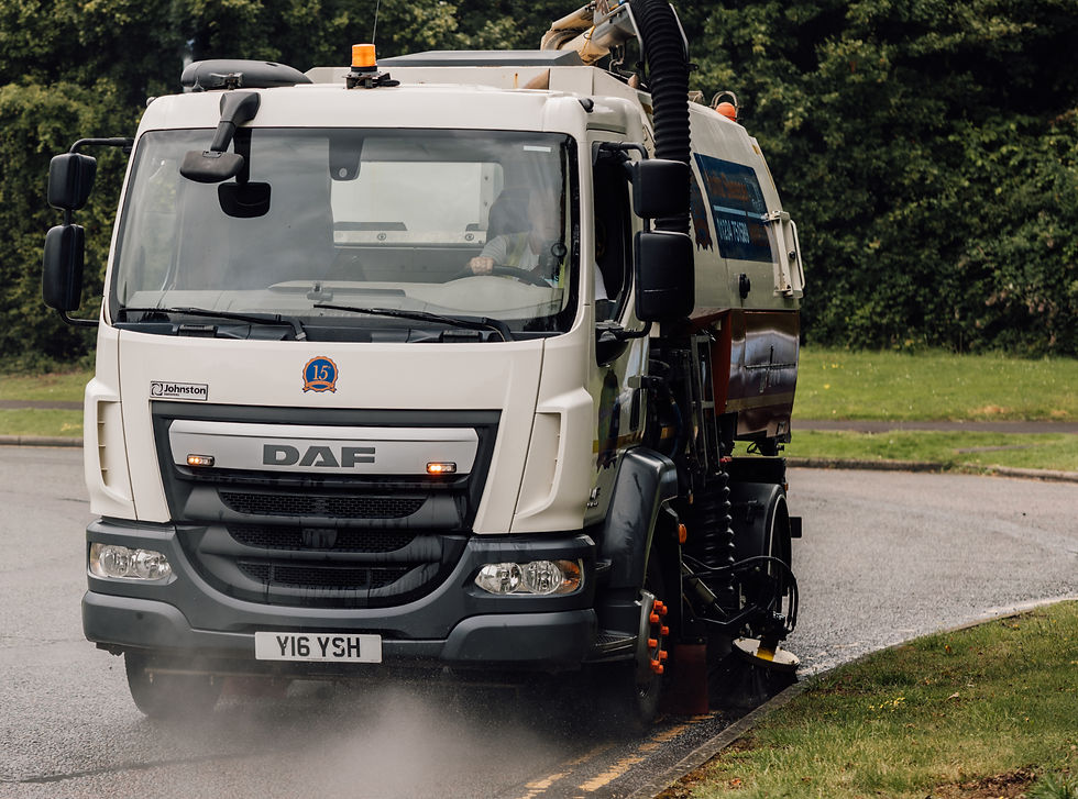 Road sweeper truck cleaning a curved road, releasing mist. White DAF truck with a 15th anniversary logo. Greenery in the background.