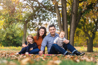 A family of 4 sits on a blanket in a park full of fall trees and leaves on the ground on a sunny fall day.
