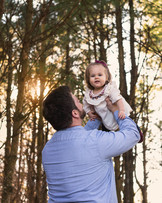 Dad flys his daughter above his head to gett giggles from her. They are in a forest area with pines trees in late fall.