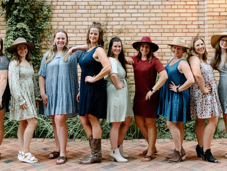 A group of nine women wearing dresses and hats stands cheerfully against a brick wall with ivy and vintage lamps. The mood is joyful and relaxed.