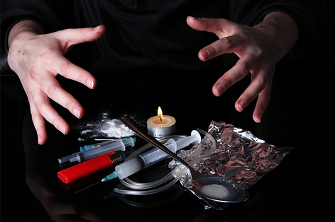 A white person's hands are stretched out over drug paraphernalia including white powder, a small lit candles, a spoon, and a syringe.
