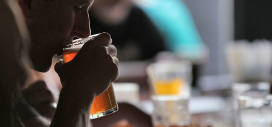 A young person is sitting in the shadows at a gathering, drinking a beer.