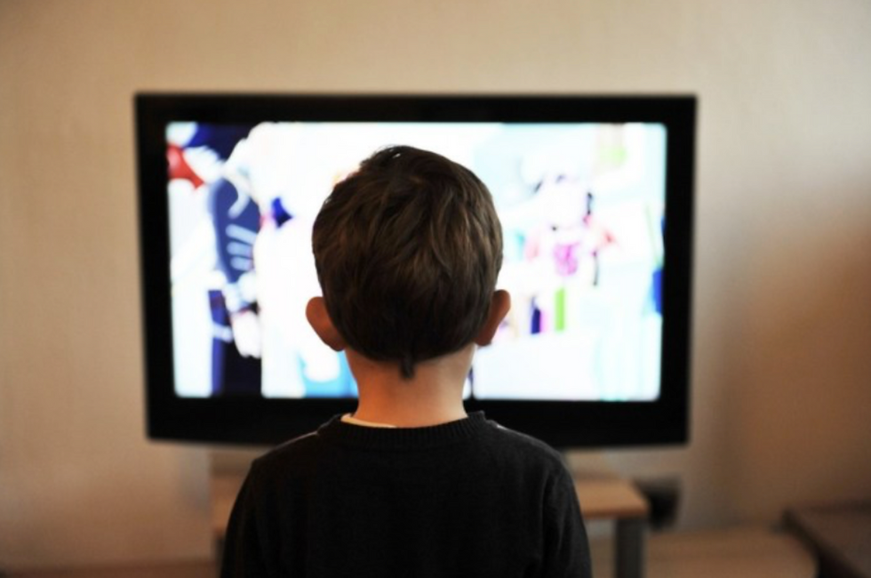 A child is facing a TV and clearly absorbing whatever he is watching. We only see the back of his head and upper torso and the content on the TV screen is blurred.