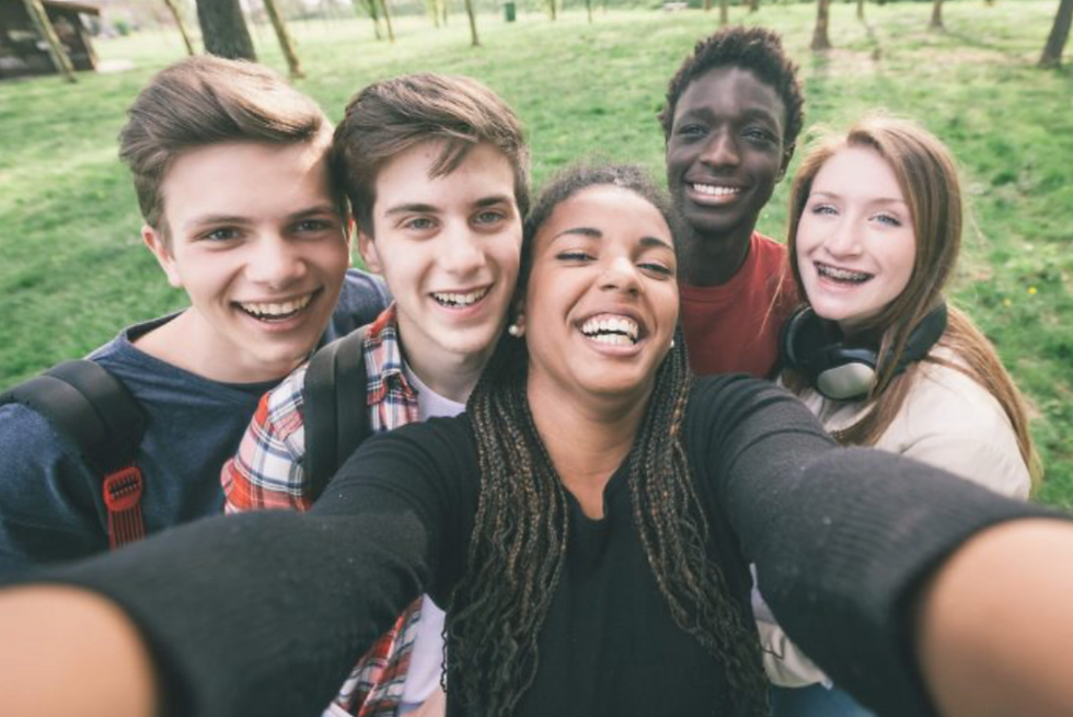 A group of teens is hanging out outside, smiling and laughing. One of them is holding a camera with two hands and taking a selfie of the group.