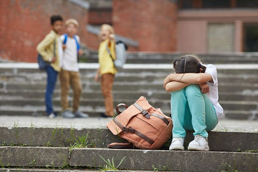 A young girl is sitting on a cement step with her head covered by her arms, her backpack on the ground next to her. In the background there are 3 other students pointing and laughing at her.