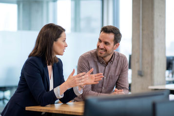 Two colleagues sitting at a table in a modern office, having an informal discussion. One person gestures while speaking, while the other listens and smiles, with desks, monitors, glass walls, and bright office lighting in the background.