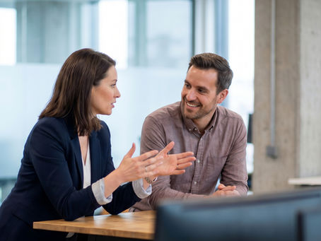 Two colleagues sitting at a table in a modern office, having an informal discussion. One person gestures while speaking, while the other listens and smiles, with desks, monitors, glass walls, and bright office lighting in the background.