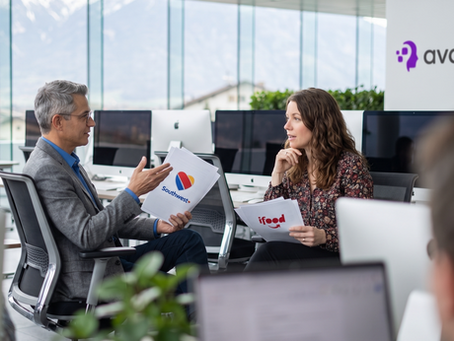 candid professional conversation between two coworkers in a modern office, medium shot, engaged in discussion with natural hand gestures, soft daylight coming from the large windows behind them, shallow depth of field, blurred foreground bokeh elements, relaxed corporate environment, authentic expressions, shot at eye level, natural composition, contemporary tech company aesthetic. One of them is holding a stack of papers with the Southwest airlines logo and the other with the ifood logo. mimic natural movements, people not aware they are being photgraphed, professional faces