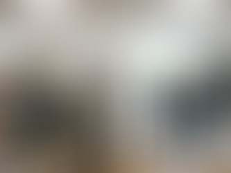 A diverse group of 8 people, dressed in casual business attire, stand in a cluttered, messy office on the left. A plain wall divides the space. On the right, a meticulously organized office features 3 people in smart suits. Bright, overhead fluorescent lighting. Midday. Spring.