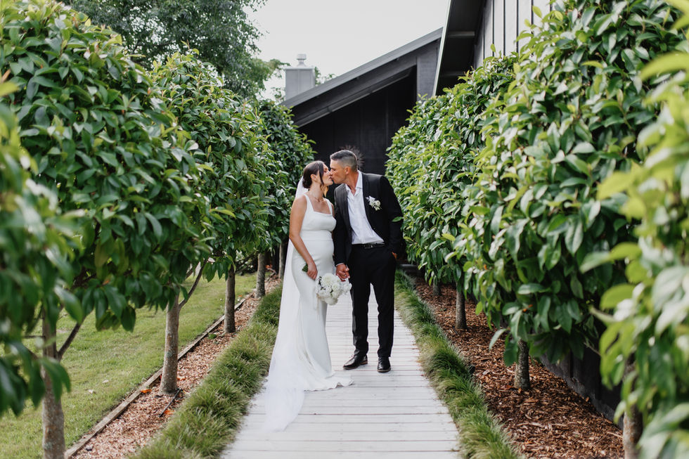Bride and groom kissing beneath a greenery tunnel while holding a modern white garden-style wedding bouquet by Sweet Stems. Romantic outdoor wedding portrait in Tauranga.