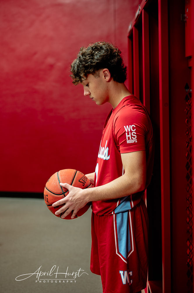 Young male basketball player in red uniform holds a ball, leaning against red lockers. "WCHS HOOPS" on sleeve. Calm, focused mood.