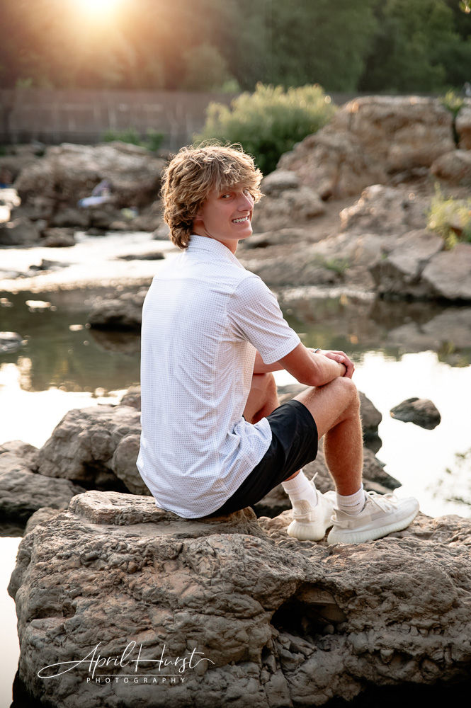 A person in a white shirt and black shorts sits on rocks by a serene water body at sunset, smiling. The scene is warm and peaceful.